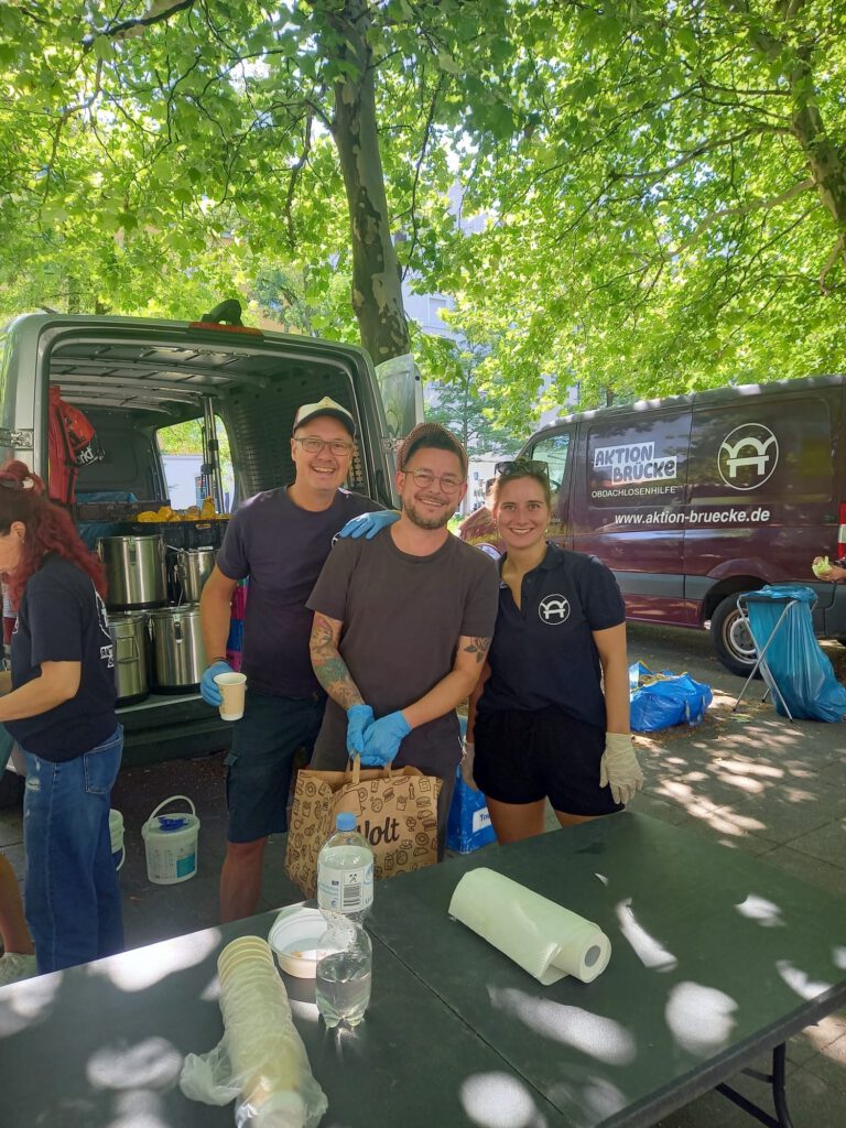 2 Männer und eine Frau vom Team Aktion Brücke stehen lächelnd vor 2 Autos mit Lebensmitteln
