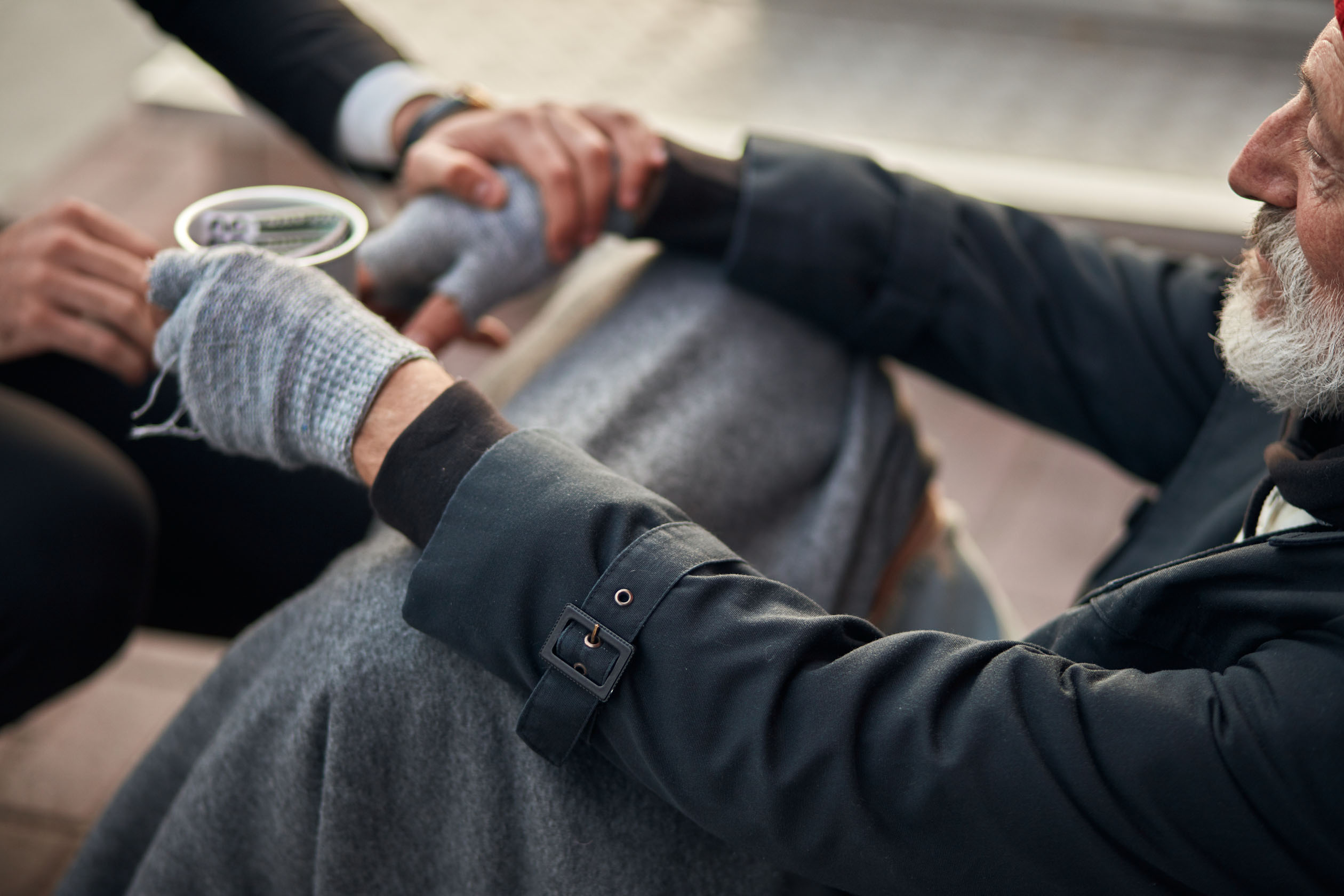 ein obdachloser älterer Mann sitzt und bittet um Spenden. eine weitere Person hat die Hand auf seine Hand gelegt