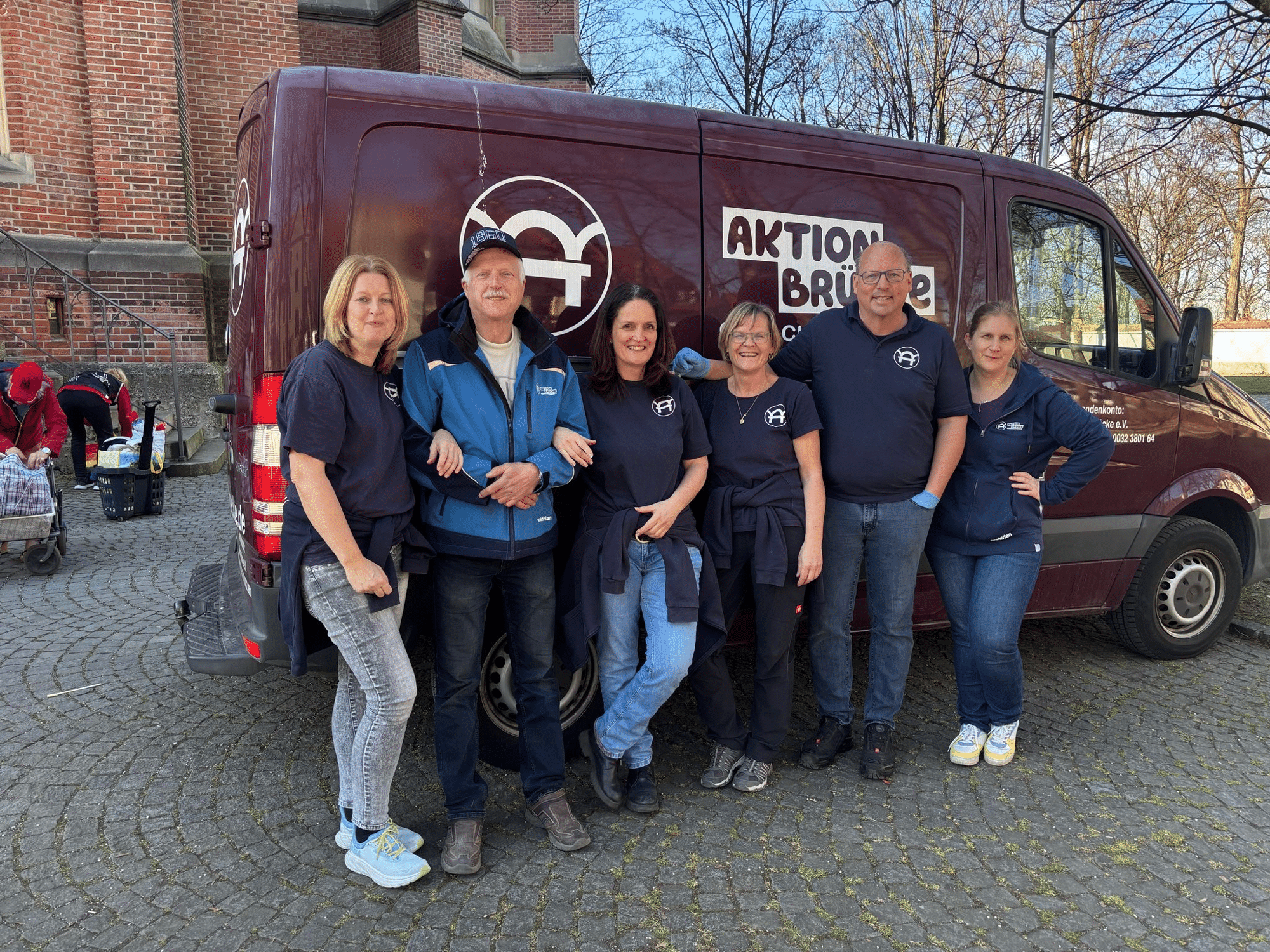 6 Personen stehen lächelnd vor einem bordeaux-farbenen Auto der Aktion Brücke